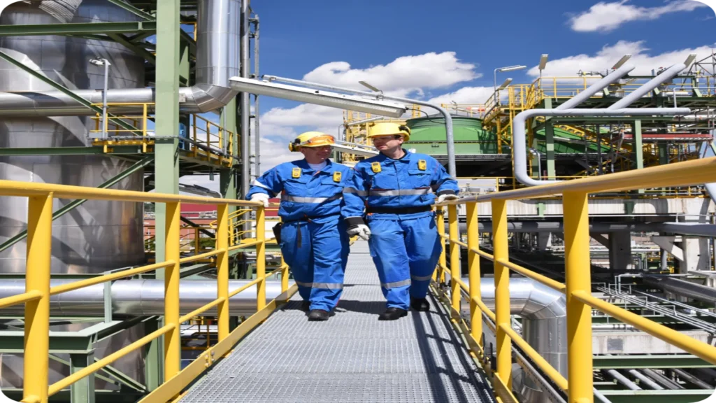 Becht workers in blue uniforms and hard hats walk along an industrial walkway with yellow railings, surrounded by piping, metal structures, and plant equipment.