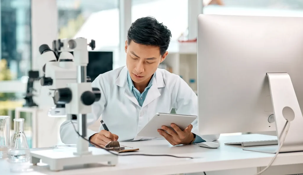 Científico joven trabajando en un laboratorio moderno, observando datos en una tablet y escribiendo en una libreta, con un microscopio y equipo de laboratorio en la mesa. La escena representa investigación científica, posiblemente relacionada con el reciclaje de plásticos.