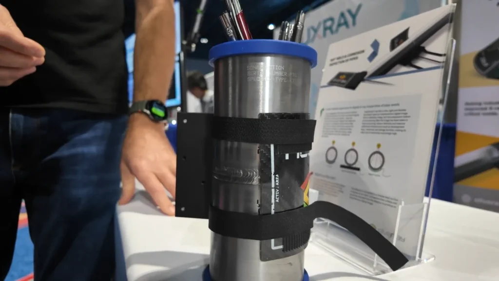 Dispositivo cilíndrico de acero inoxidable con correas de velcro negras sujetando un detector digital de rayos X de Silveray, mostrado sobre una mesa de exhibición durante la convención ASNT 2025, como ejemplo de innovación en la digitalización de soldaduras.