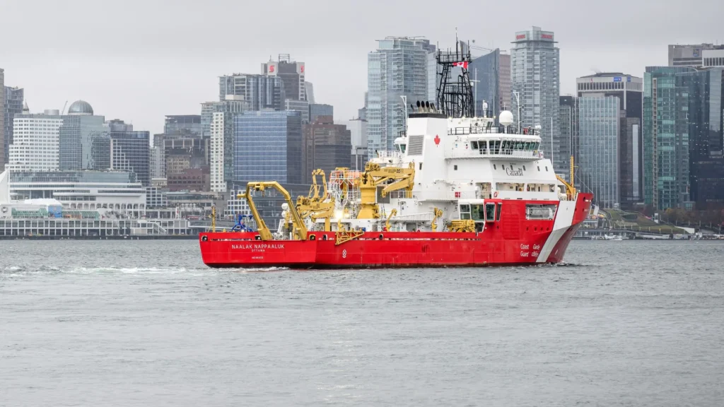 Buque científico CCGS Naalak Nappaaluk navegando frente a la costa de Vancouver con el skyline urbano de fondo.