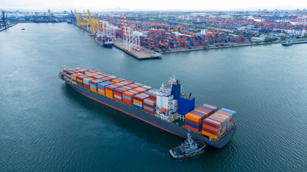 Vista aérea de terminales marítimos con un buque portacontenedores maniobrando junto a un puerto equipado con grúas, patios de contenedores e infraestructura logística.